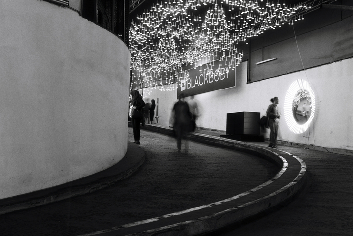 Art exhibition on a carpark ramp with dangling lights, people and a mirror