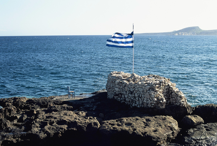 A greek flag and a chair on a rocky outcrop in the sea
